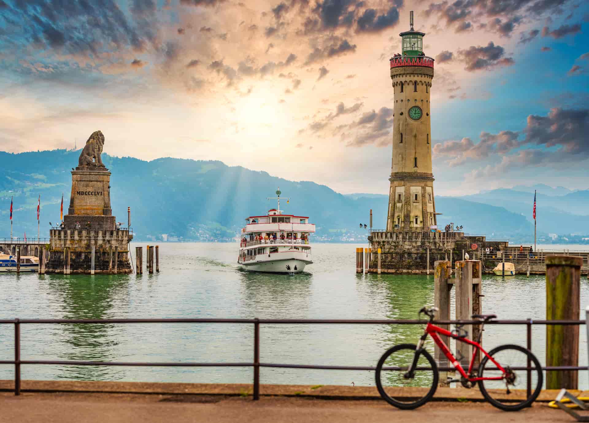Lion statue and lighthouse flanking a boat on Lake Constance with mountains in Lindau, Germany.