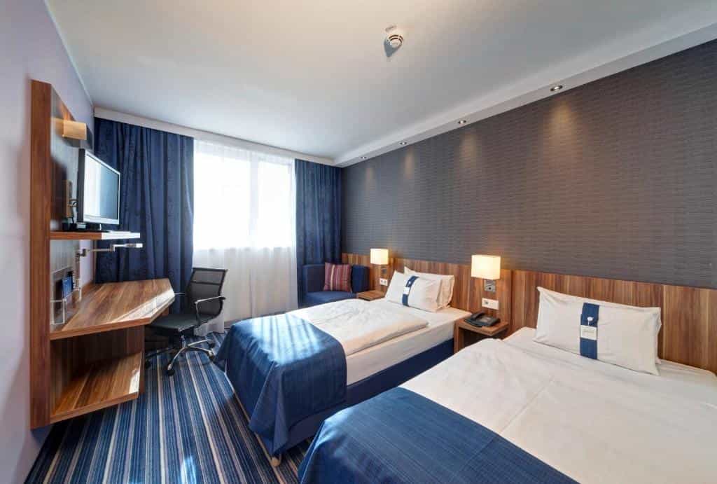 Twin beds in a modern hotel room with blue striped carpet and wooden desk area in Augsburg.