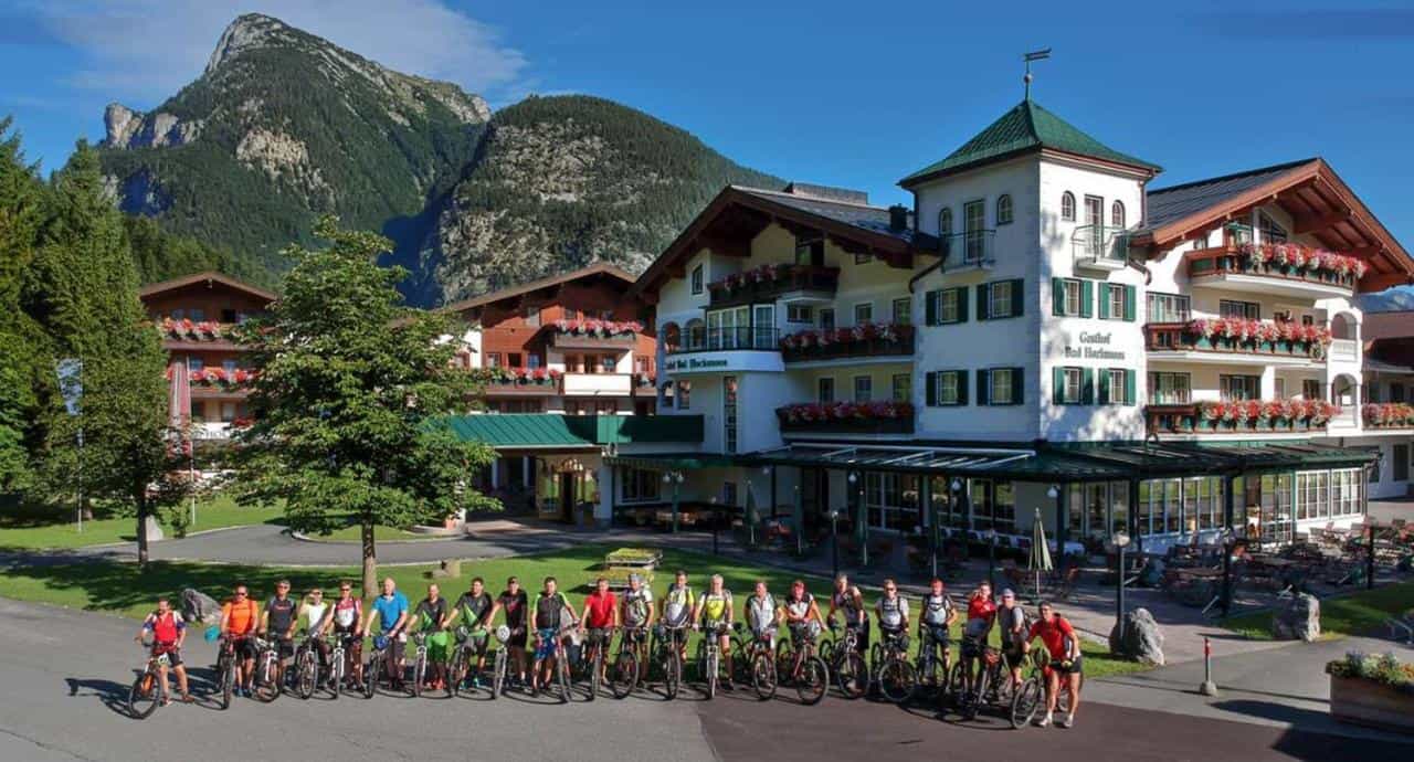 Group of mountain bikers outside a traditional alpine hotel with large mountains in background.