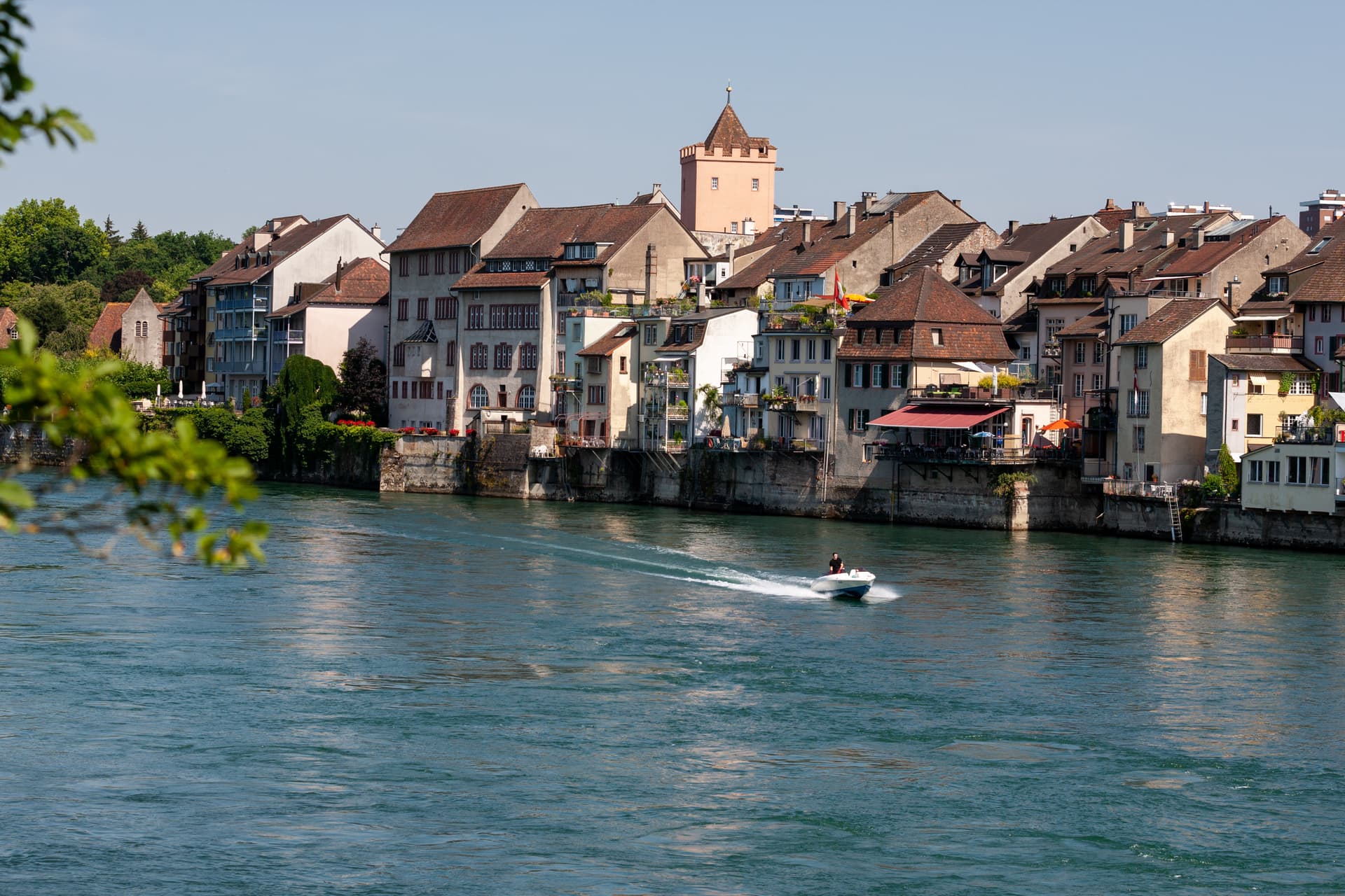 Motorboat speeding on river past historic buildings in Rheinfelden with leafy foreground.