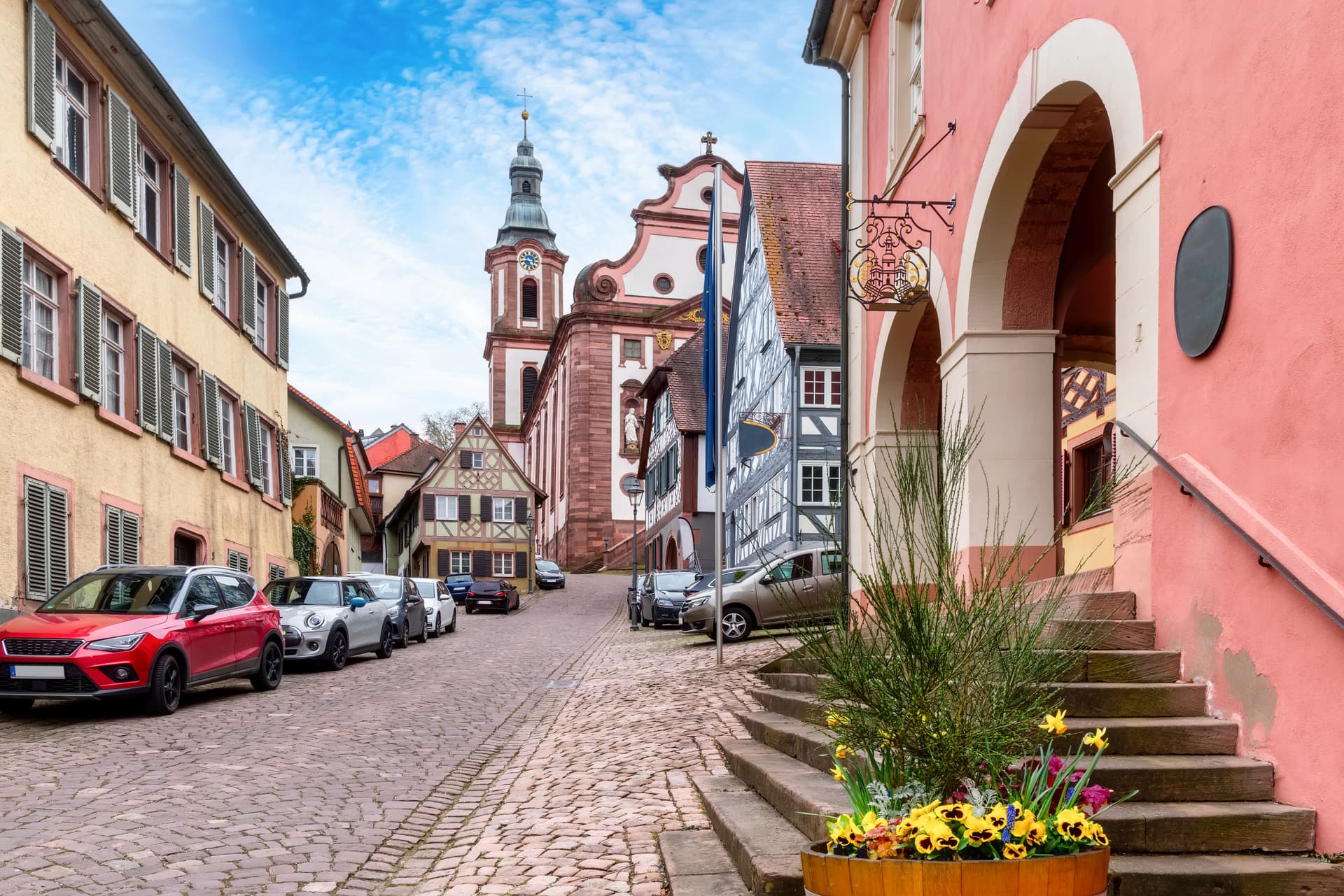Cobblestone street in Ettenheim leading to a church tower, with parked cars and historic buildings.