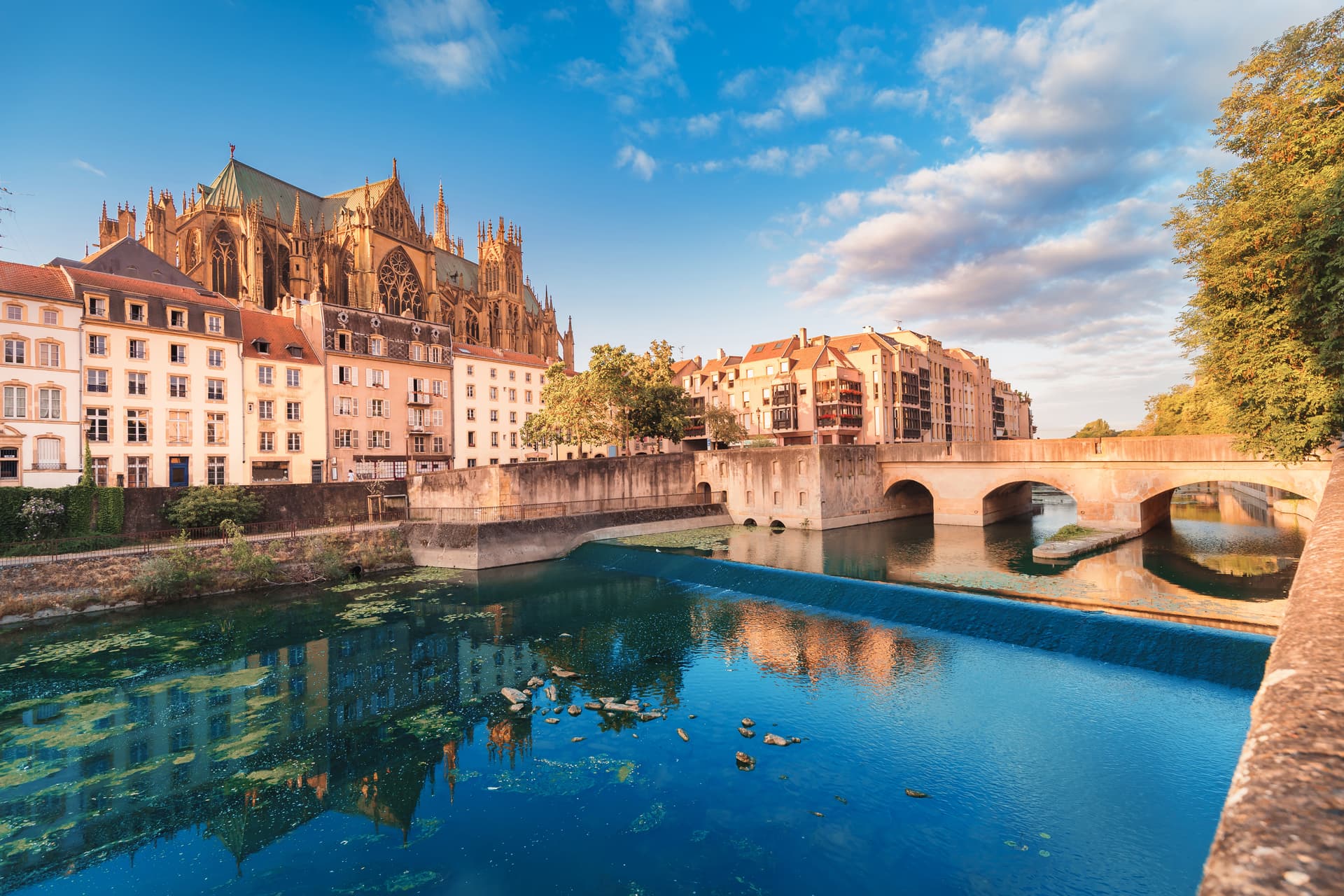 Metz waterfront with Saint-Étienne Cathedral, river, and stone bridge under blue sky.