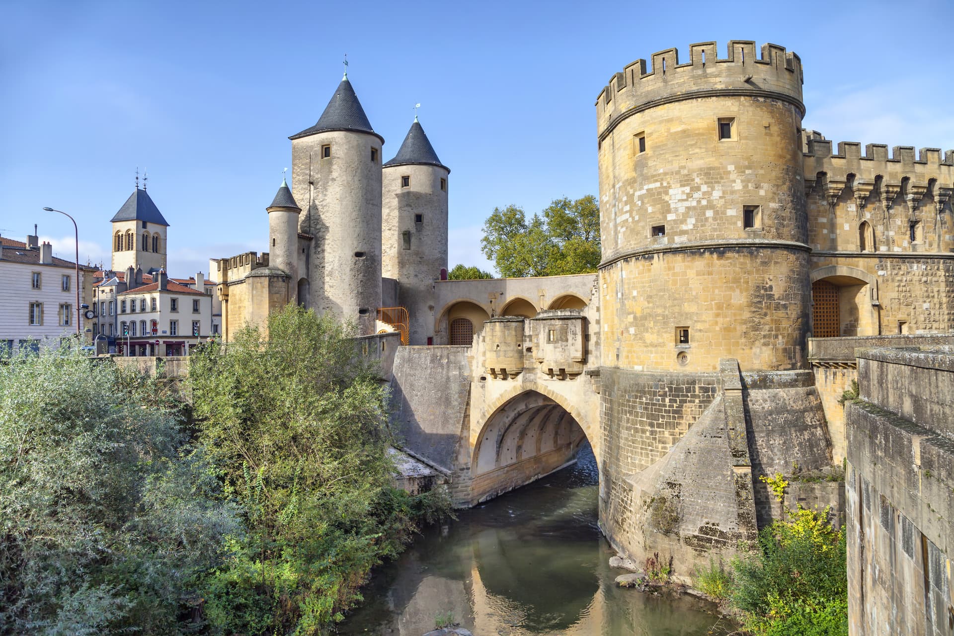 Fortified medieval gatehouse with towers over a river, with town buildings in the background.