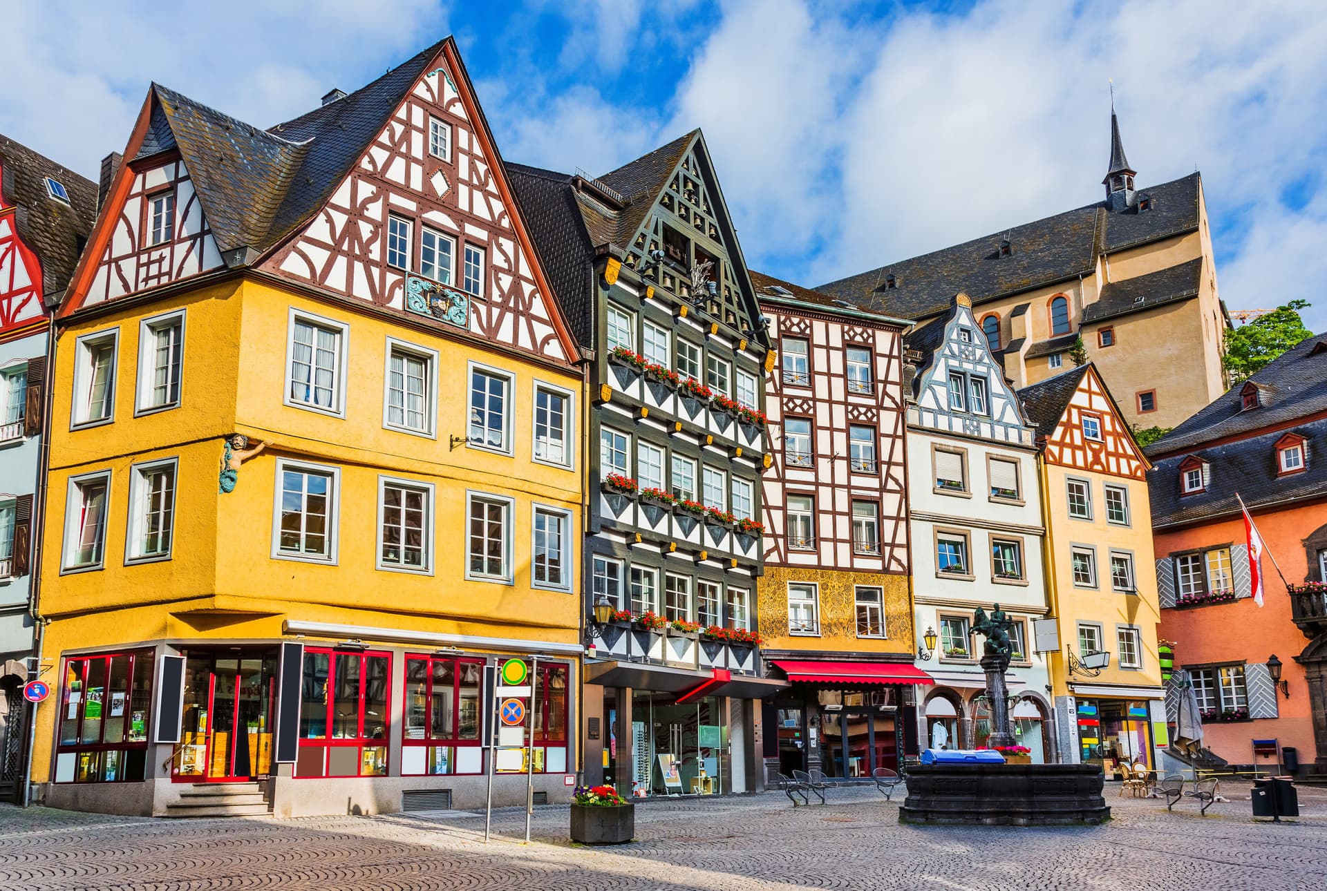 Half-timbered buildings line Cochem square with a fountain and church steeple under blue sky.