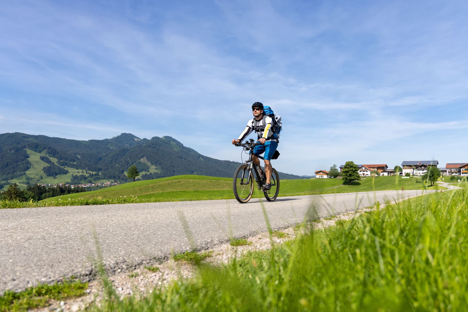Cyclist with backpack riding on paved road through green hills toward Bavarian village and mountains.