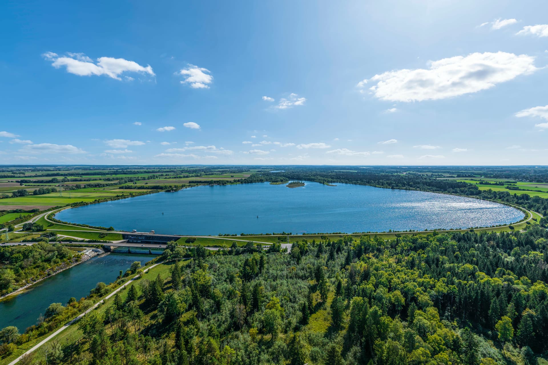 Aerial view of Lech Lake with dam structure, green forests, and surrounding agricultural fields under a blue sky.