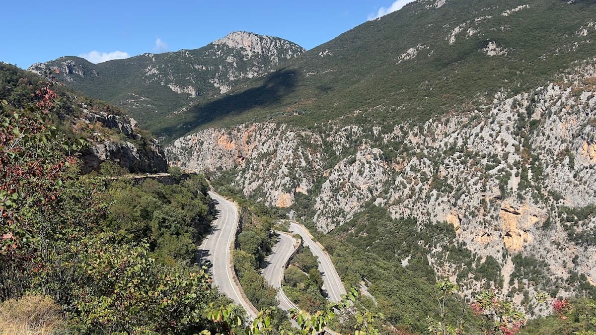 Winding mountain road traversing steep, rocky slopes covered in green vegetation under a blue sky.