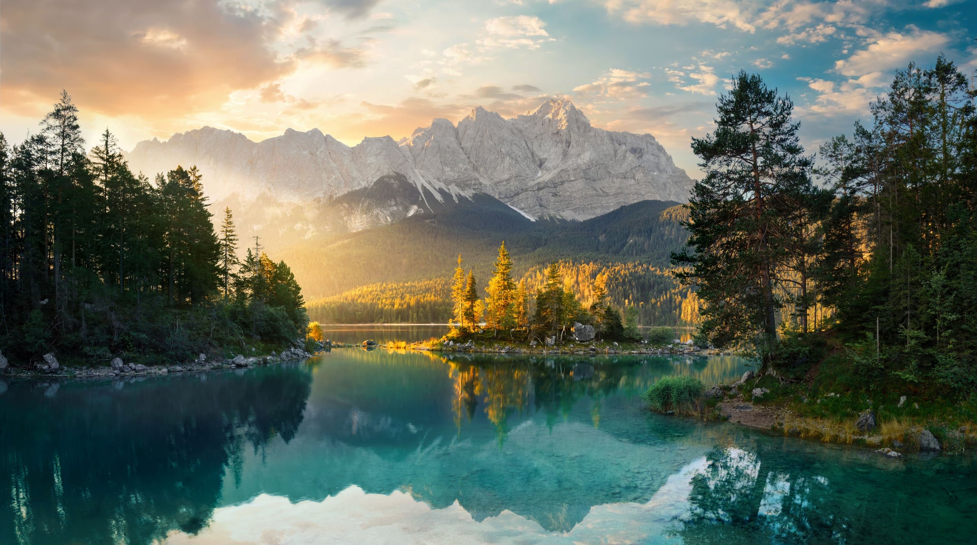 Alpine lake with turquoise water reflecting sunlit mountains and pine forests at sunrise or sunset.
