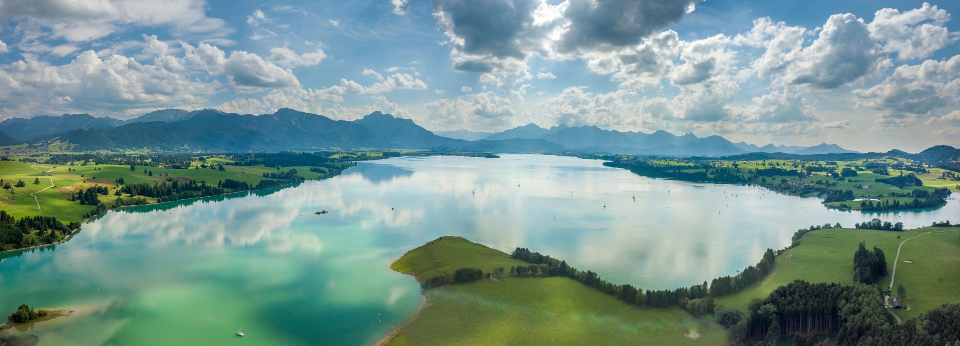 Aerial view of Forggensee lake with turquoise water, green shores, and distant mountains under a cloudy blue sky.