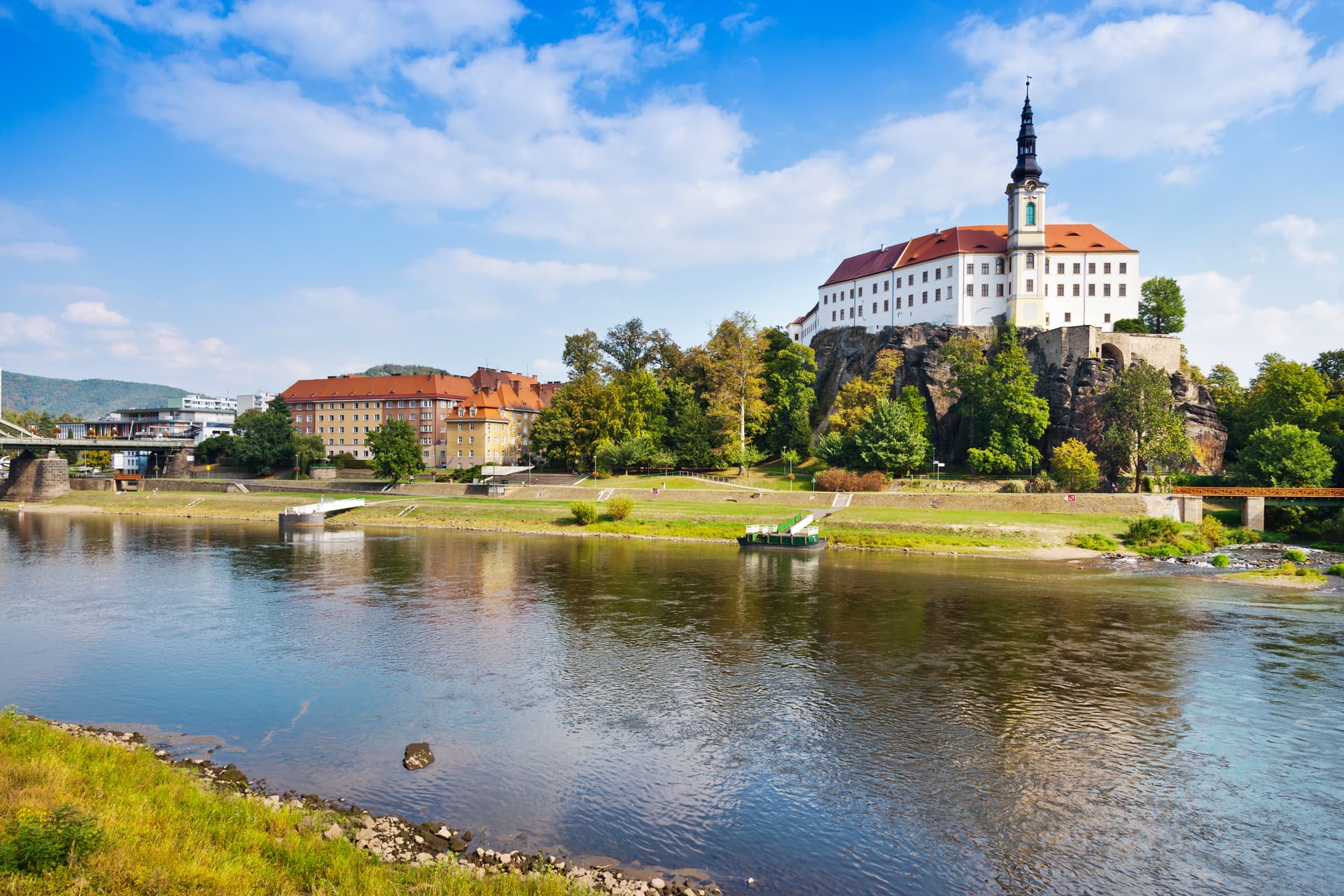 Dečín castle on a cliff above a river with a bridge and town buildings.