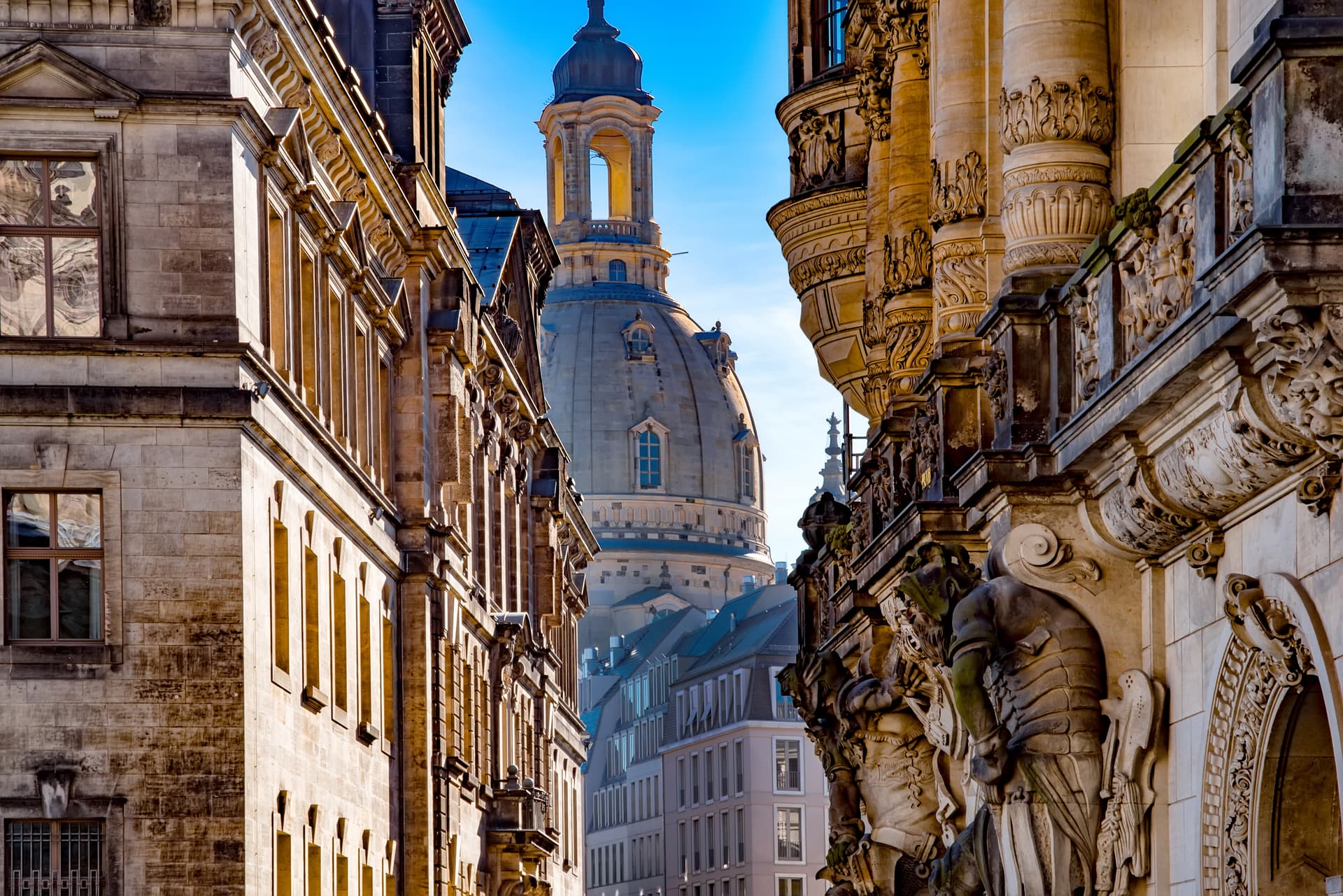 Baroque architecture and dome visible between buildings on Dresden alley under clear blue sky.