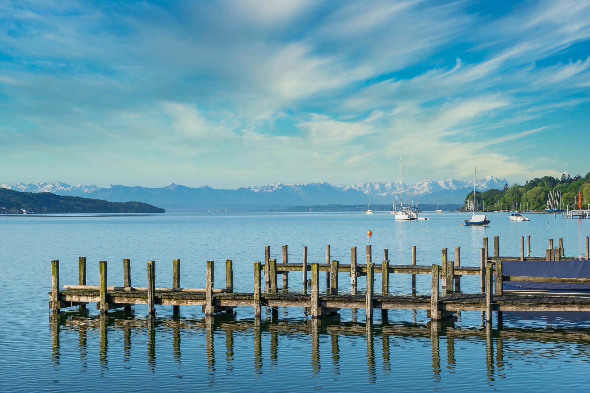 Wooden dock pilings reflected in Lake Starnberg with sailboats and snow-capped Alps.