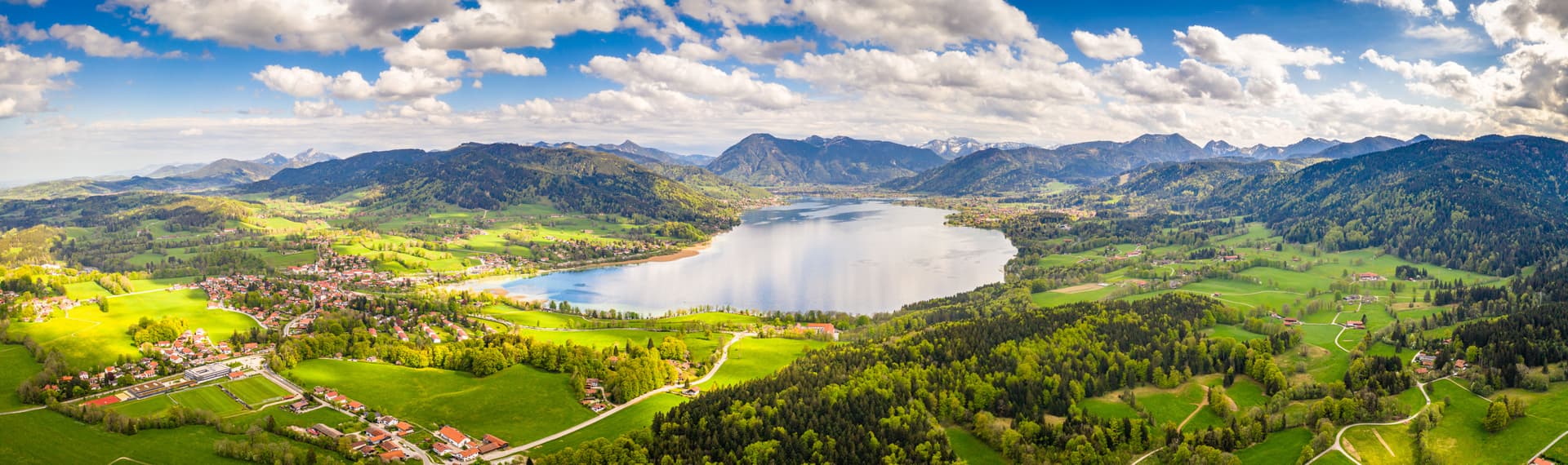 Aerial view of Lake Tegernsee surrounded by green hills, forests, and distant snow-capped mountains under a blue sky.