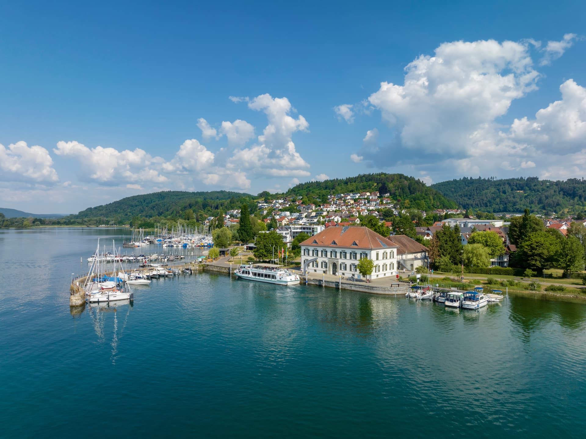 Aerial view of Bodman-Ludwigshafen harbor with sailboats, old customs house, and green hills.